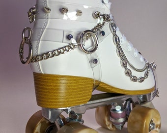 Peut inclure: Patin à roulettes blanc avec des éléments métalliques argentés, des roues marron et un talon en bois. Le patin est orné de lanières transparentes avec des pointes argentées et une chaîne. Un charme de crâne est visible sur le côté.