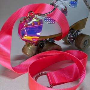 May include: A pair of white roller skates with pink laces and a pink and silver strap. The skates have a holographic, spiked toe stop and a pink strap with a silver ring.