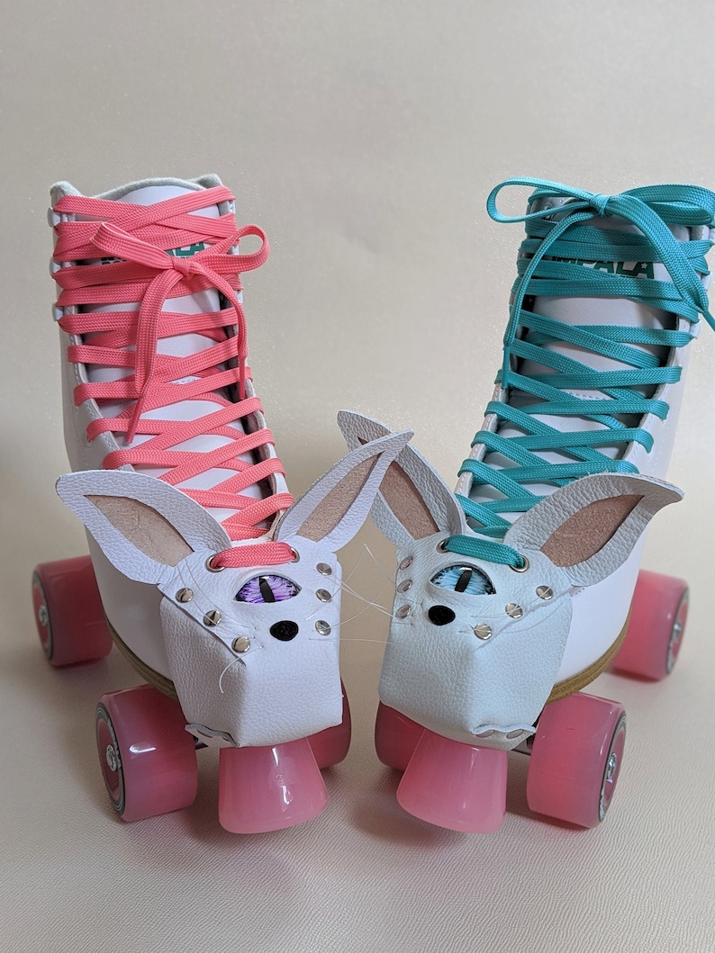 May include: A pair of white roller skates with pink and teal laces. Each skate has a white leather bunny face attached, complete with ears, eyes, and nose. The wheels are pink.