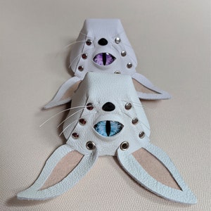 May include: Two white leather bunny masks with detailed eye designs. Each mask has a black nose, whiskers, and silver rivets. One mask has purple eyes, and the other has blue eyes, with pink inner ears.