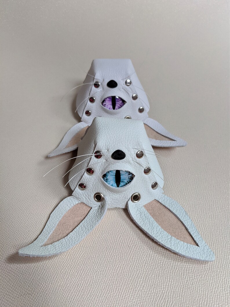 May include: Two white leather bunny masks with detailed eye designs. Each mask has a black nose, whiskers, and silver rivets. One mask has purple eyes, and the other has blue eyes, with pink inner ears.