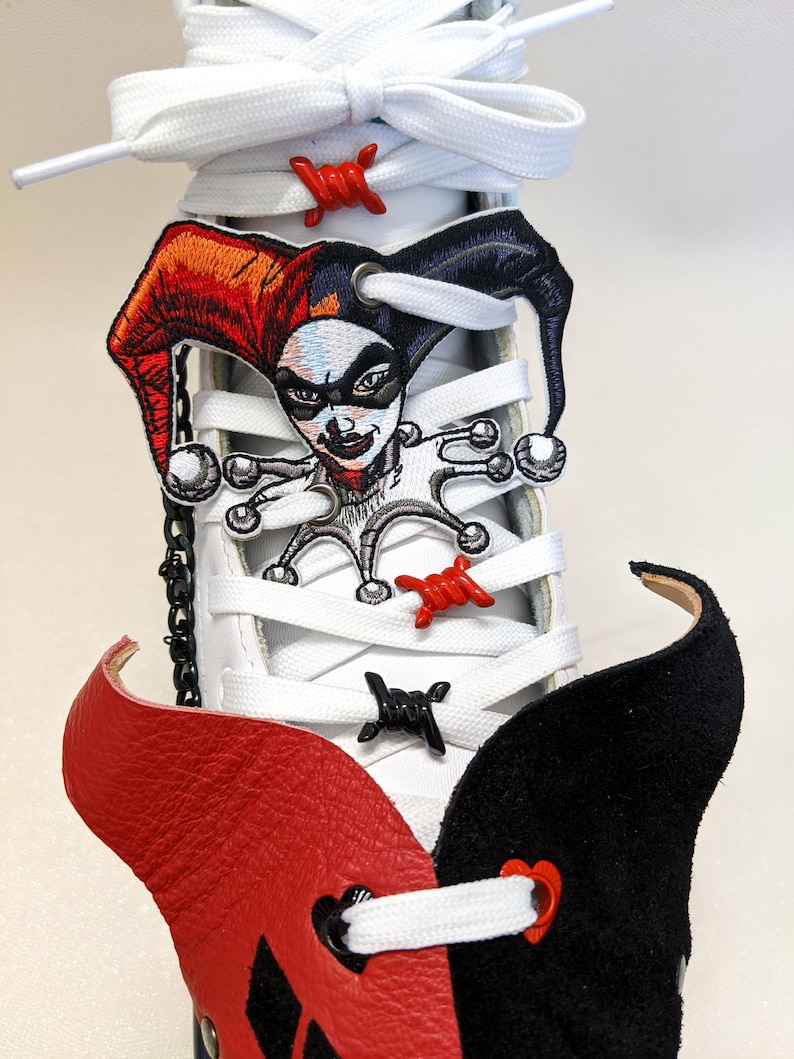 May include: White high-top sneakers with red and black leather accents. The laces are white and feature red and black barbed wire charms. A Harley Quinn character patch is attached to the laces.