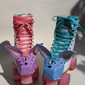 May include: Two roller skates with white boots, pink wheels, and colorful laces. Each skate has a leather bunny face in purple and blue, with a third eye design. The laces are coral and teal.