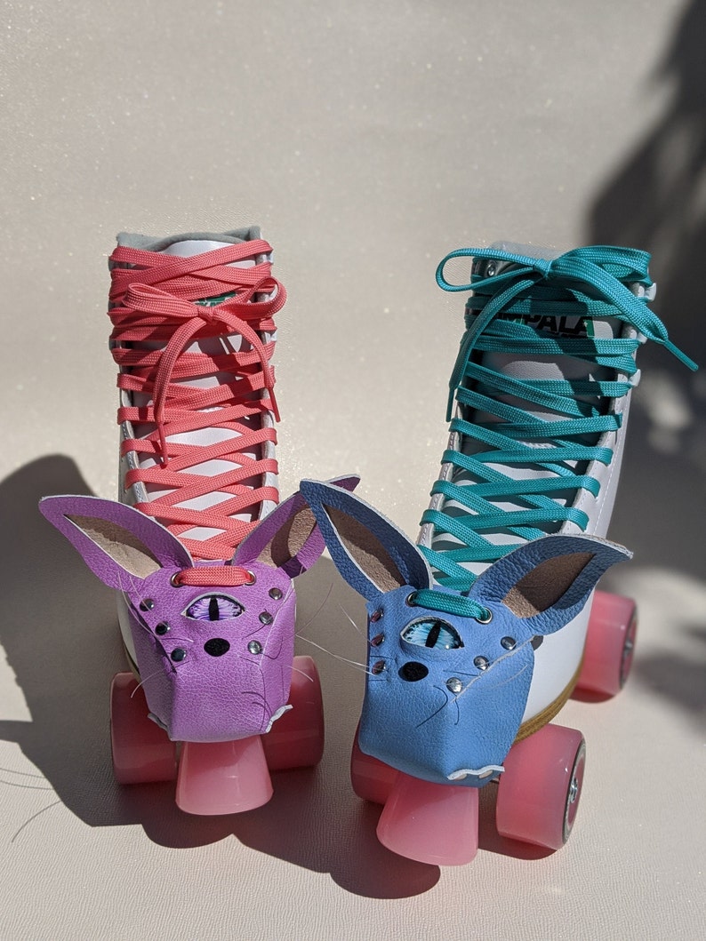 May include: Two roller skates with white boots, pink wheels, and colorful laces. Each skate has a leather bunny face in purple and blue, with a third eye design. The laces are coral and teal.