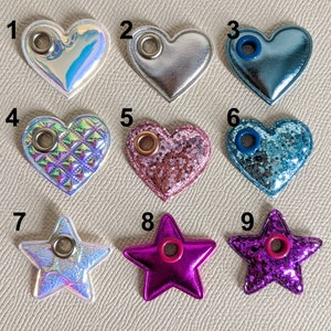May include: Nine heart and star-shaped embellishments with eyelets. The embellishments are in various colors and textures, including iridescent, glitter, and quilted.
