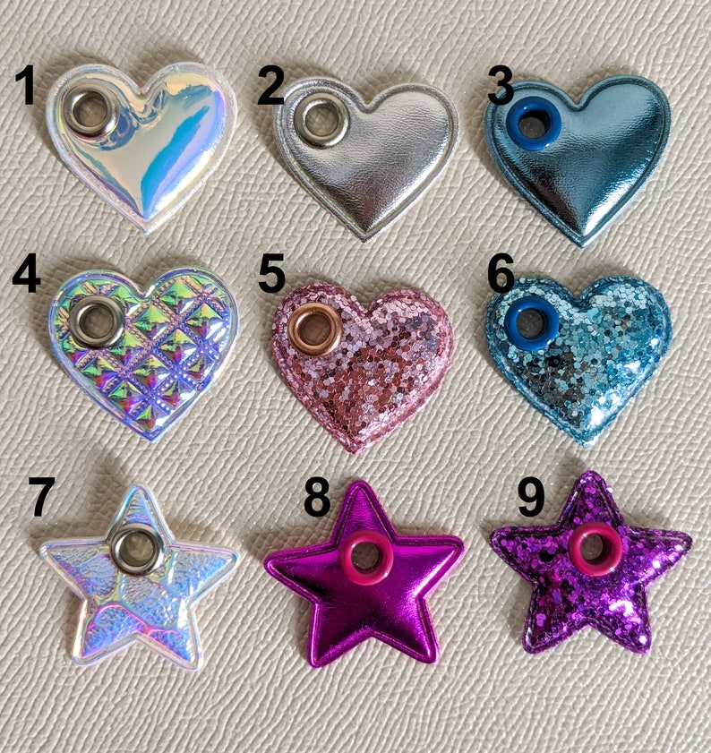 May include: Nine heart and star-shaped embellishments with eyelets. The embellishments are in various colors and textures, including iridescent, glitter, and quilted.