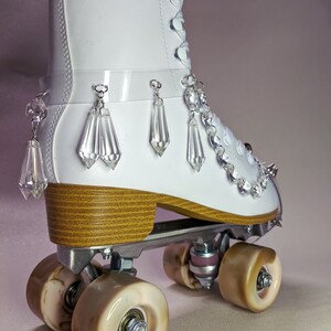 May include: White roller skates with a clear plastic strap and crystal embellishments. The skates have a wooden platform and brown wheels.