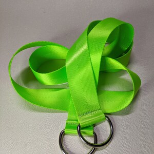 Puede incluir: Una correa de nylon verde brillante con dos anillos en D de metal plateado. La correa est&aacute; doblada y colocada sobre una superficie blanca.