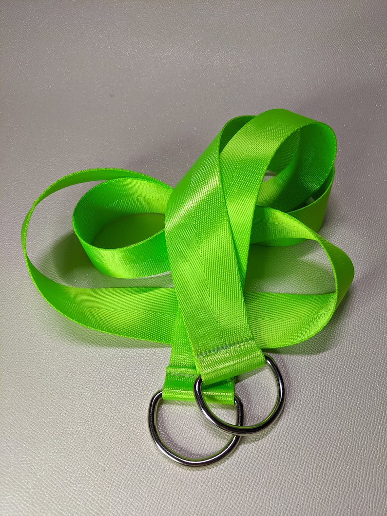 Puede incluir: Una correa de nylon verde brillante con dos anillos en D de metal plateado. La correa est&aacute; doblada y colocada sobre una superficie blanca.