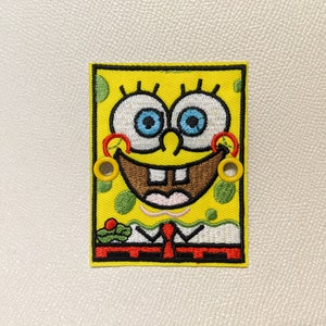 May include: Yellow embroidered patch of a cartoon character with a big smile, blue eyes, and a red tie. The character has a yellow background with green and white details.