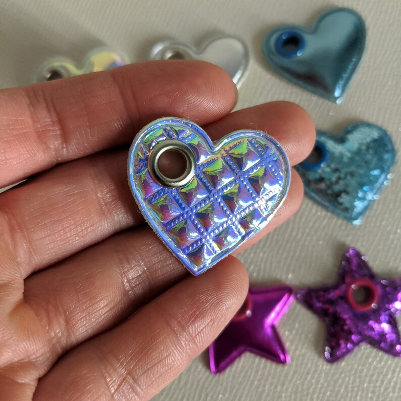 May include: A light blue iridescent heart-shaped charm with a silver metal eyelet. The charm has a quilted pattern and is made of a shiny material.