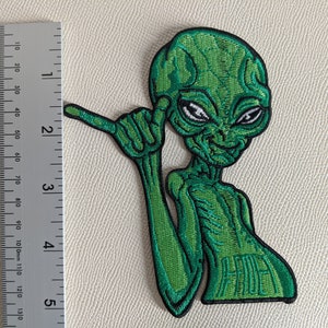 May include: Green embroidered iron-on patch of an alien with a mischievous expression, giving a shaka sign with its right hand. The alien has a large head and large eyes. The text "HAWAII" is embroidered on the bottom of the patch.