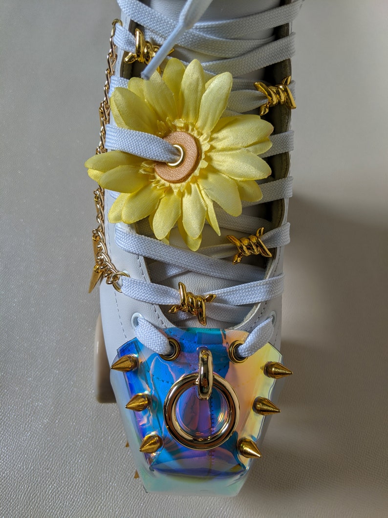 May include: White roller skate with gold chain, barbed wire, and a yellow flower on the laces. The toe stop is a gold ring with spikes.