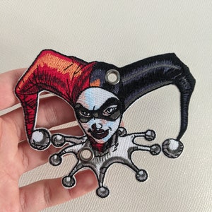 May include: Embroidered patch featuring Harley Quinn, a comic book character, with a red and black jester hat and a white and gray jester costume. The patch has two metal eyelets.