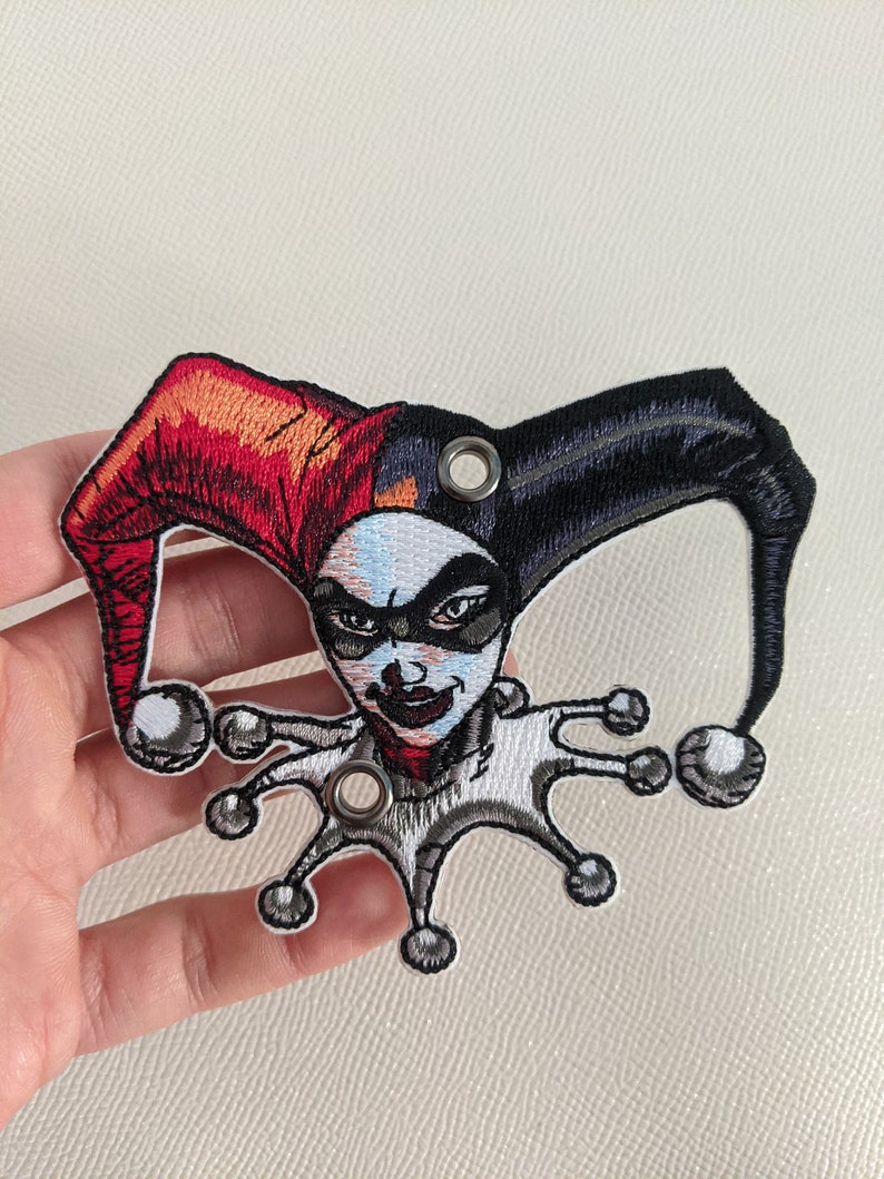 May include: Embroidered patch featuring Harley Quinn, a comic book character, with a red and black jester hat and a white and gray jester costume. The patch has two metal eyelets.