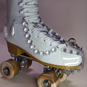 May include: White roller skate with clear plastic toe guard and crystal embellishments. The skate has brown wheels and a white leather boot.