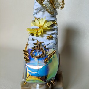 May include: White roller skate with a gold butterfly, yellow flower, and gold chain charm. The skate has a iridescent, rainbow-colored toe stop with gold spikes.