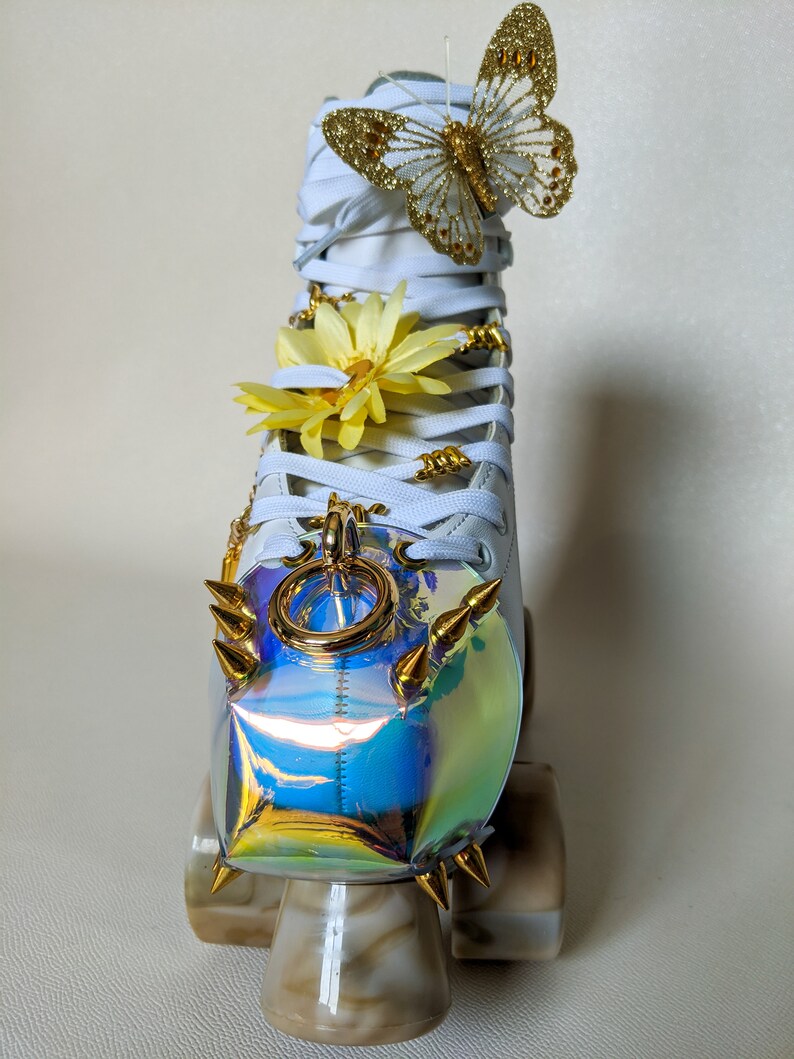 May include: White roller skate with a gold butterfly, yellow flower, and gold chain charm. The skate has a iridescent, rainbow-colored toe stop with gold spikes.