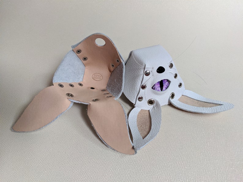 May include: White leather bunny mask with purple eye detail and silver rivets. The mask is open, revealing the interior construction and a "HAND MADE" stamp. The mask has long ears and is designed to be worn.