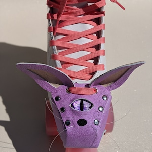 May include: White roller skate with coral laces and pink wheels. A purple leather cat-shaped accessory is attached to the front, featuring an eye design and silver studs. The cat accessory has long whiskers.