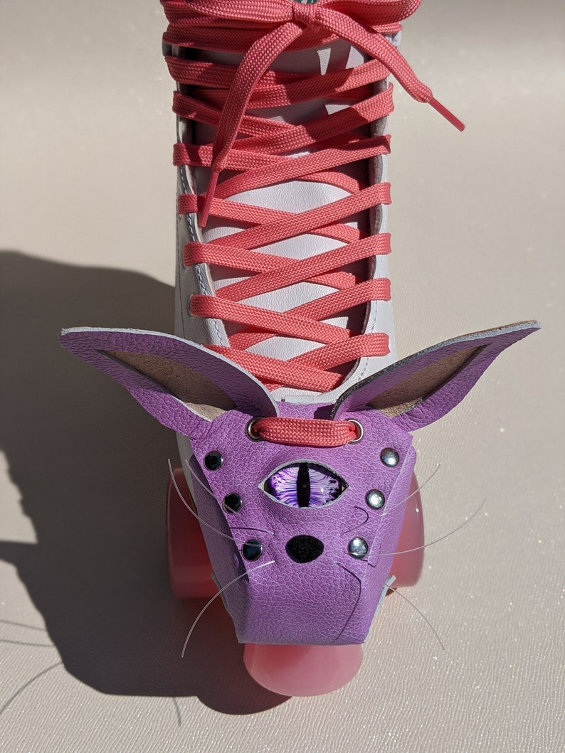 May include: White roller skate with coral laces and pink wheels. A purple leather cat-shaped accessory is attached to the front, featuring an eye design and silver studs. The cat accessory has long whiskers.