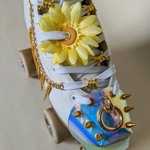 May include: White roller skates with gold accents. The skates have a yellow flower charm, gold chain, and gold spikes. The toe stop is a clear iridescent plastic with gold spikes.