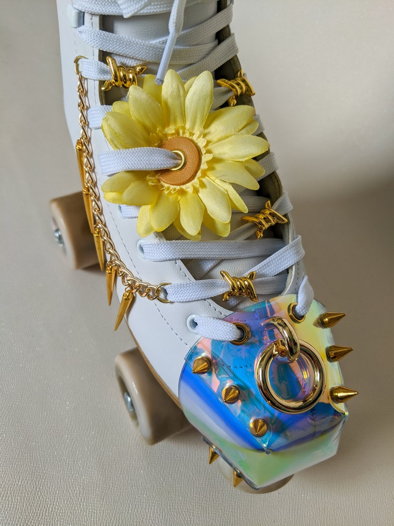 May include: White roller skates with gold accents. The skates have a yellow flower charm, gold chain, and gold spikes. The toe stop is a clear iridescent plastic with gold spikes.
