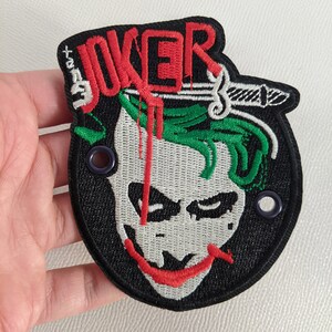 Peut inclure: Patch brodé noir et blanc avec un visage de Joker vert et rouge. Le mot "JOKER" est brodé en rouge au-dessus du visage. Le patch a deux œillets sur les côtés.