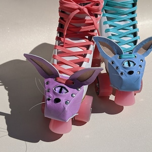 May include: Pair of roller skates with custom bunny ear covers. One skate has a purple cover with a single eye design, the other has a blue cover. Both skates have pink wheels and white boots with coral and teal laces.