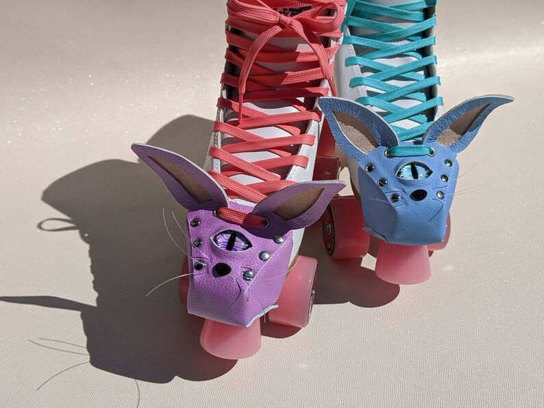 May include: Pair of roller skates with custom bunny ear covers. One skate has a purple cover with a single eye design, the other has a blue cover. Both skates have pink wheels and white boots with coral and teal laces.