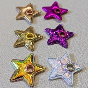 May include: Six star-shaped embellishments with metal eyelets. The stars are made of different materials, including glitter, iridescent fabric, and metallic leather. The colors include gold, pink, purple, and white.
