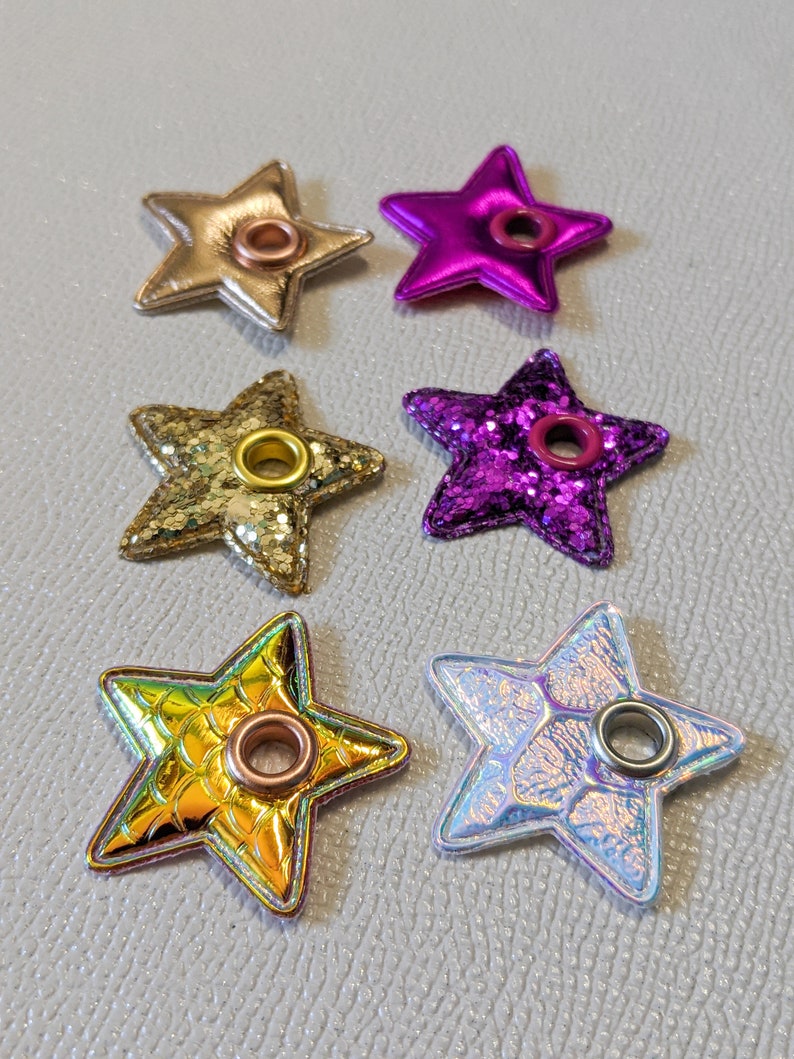 May include: Six star-shaped embellishments with metal eyelets. The stars are made of different materials, including glitter, iridescent fabric, and metallic leather. The colors include gold, pink, purple, and white.
