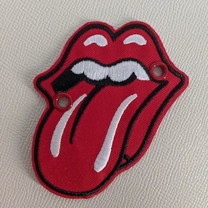 May include: A red embroidered patch in the shape of a tongue and lips, outlined in black. The tongue has white stitching and two red grommets. The patch is on a textured, off-white surface.