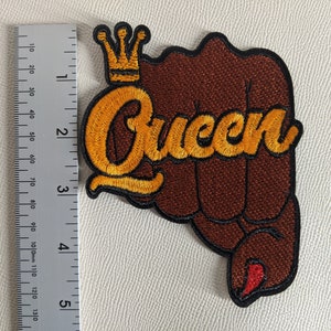 Pode incluir: Um punho marrom com uma coroa dourada e a palavra "Queen" bordada em ouro. O punho está levantado no ar.