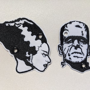 May include: Two black and white embroidered patches of Frankenstein's monster and his bride. The patches have black stitching and white faces. Each patch has three black eyelets.