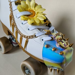 May include: White roller skate with gold accents, a yellow flower, and a clear iridescent toe stop with gold studs. The skate has a gold chain and spikes.