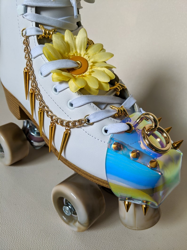 May include: White roller skate with gold accents, a yellow flower, and a clear iridescent toe stop with gold studs. The skate has a gold chain and spikes.