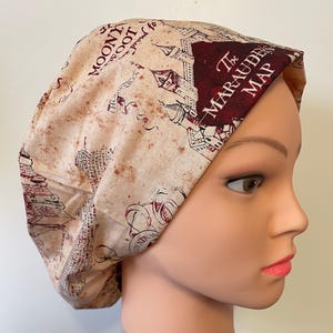 May include: A beige and burgundy patterned scrub cap featuring a map design with text that reads "The Marauder's Map" and "Moony, Wormtail, Padfoot, and Prongs". The cap is made of fabric and is designed to cover the head.