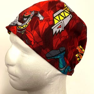 May include: A red and maroon patterned scrub cap featuring Power Rangers characters. The cap has a repeating pattern of Power Rangers figures and geometric shapes. The cap is made of a soft, lightweight fabric.