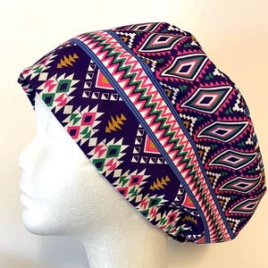 May include: A patterned scrub cap in shades of purple, pink, green, and yellow. The cap features a geometric design with diamond and zigzag patterns. The cap is made of fabric and is designed to be worn on the head.