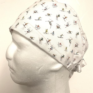 May include: White surgical scrub cap featuring a repeating pattern of skiers in action. The skiers are illustrated in a range of colours. The cap is made from a soft, lightweight material, suitable for medical or culinary use.