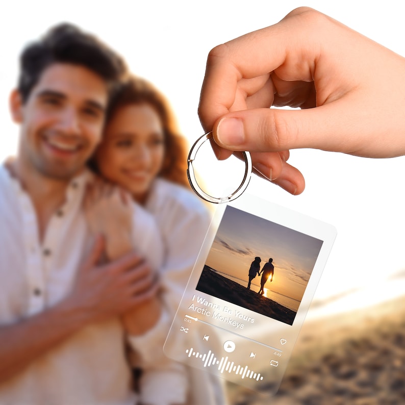 May include: A clear acrylic keychain with a silver ring, featuring a photo of a couple holding hands at sunset. The keychain displays a music player interface with the song title "I Wanna Be Yours" by Arctic Monkeys.