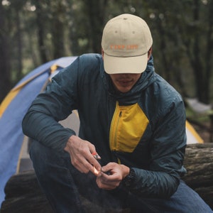 May include: A person wearing a beige baseball cap with the text "CAMP LIFE" in orange. The person is sitting on a log in a forest setting.