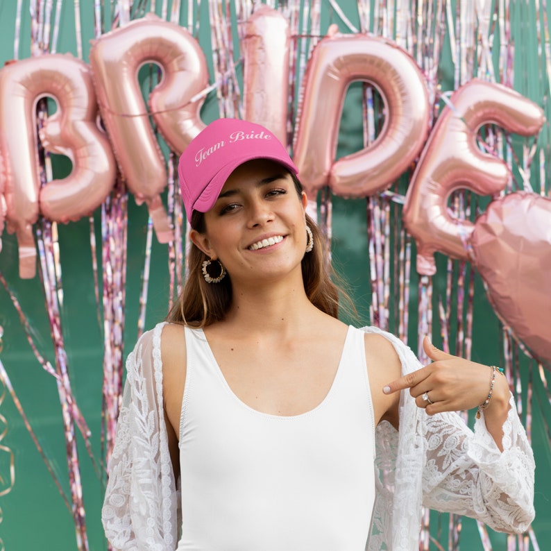 May include: A woman wearing a pink baseball cap with the text "Team Bride" in white lettering. She is smiling and pointing to her white tank top. The background is a green wall with pink balloons that spell out "BRIDE".