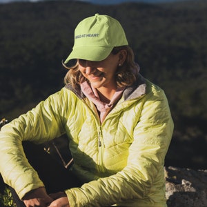 May include: A woman wearing a bright yellow jacket and a lime green baseball cap with the text "WILD AT HEART" embroidered on the front. She is sitting on a rock with a green and brown background.