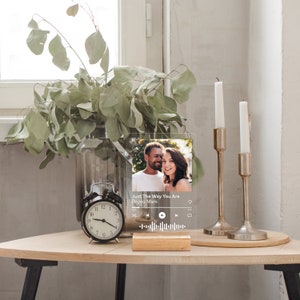 May include: A clear acrylic plaque with a photo of a couple and the text "Just The Way You Are Bruno Mars." The plaque is displayed on a wooden base. A black alarm clock, candles, and a vase with greenery are also on the table.