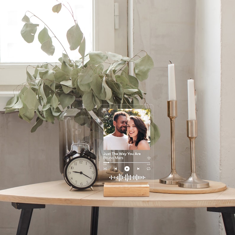 May include: A clear acrylic plaque with a photo of a couple and the text "Just The Way You Are Bruno Mars." The plaque is displayed on a wooden base. A black alarm clock, candles, and a vase with greenery are also on the table.