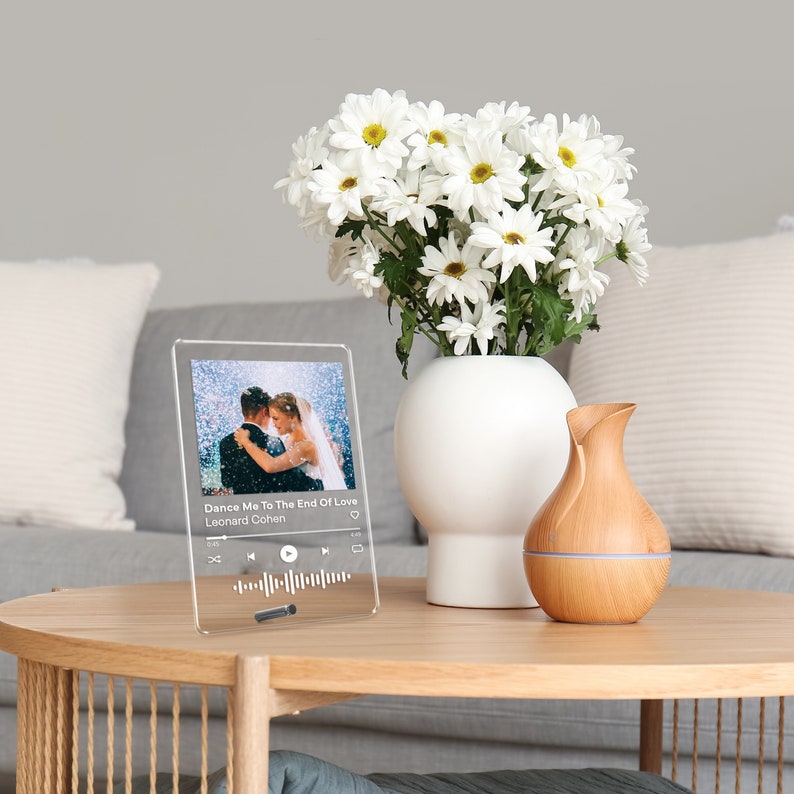 May include: A clear acrylic plaque displays a wedding photo with the text "Dance Me To The End Of Love" by Leonard Cohen. The plaque sits on a wooden table next to a white vase with white flowers and a wooden essential oil diffuser.