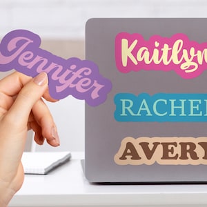 May include: A hand holds a purple sticker with the name "Jennifer" in a script font. Other stickers on a laptop read "Kaitlyn", "Rachel", and "Avery". The stickers are in various colors and fonts, suitable for personalizing laptops or other items.