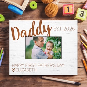 May include: White wooden photo frame with the word "Daddy" in brown script, "EST. 2026", and a photo of a man holding a baby. The frame also includes the text "HAPPY FIRST FATHER'S DAY" and "ELIZABETH".